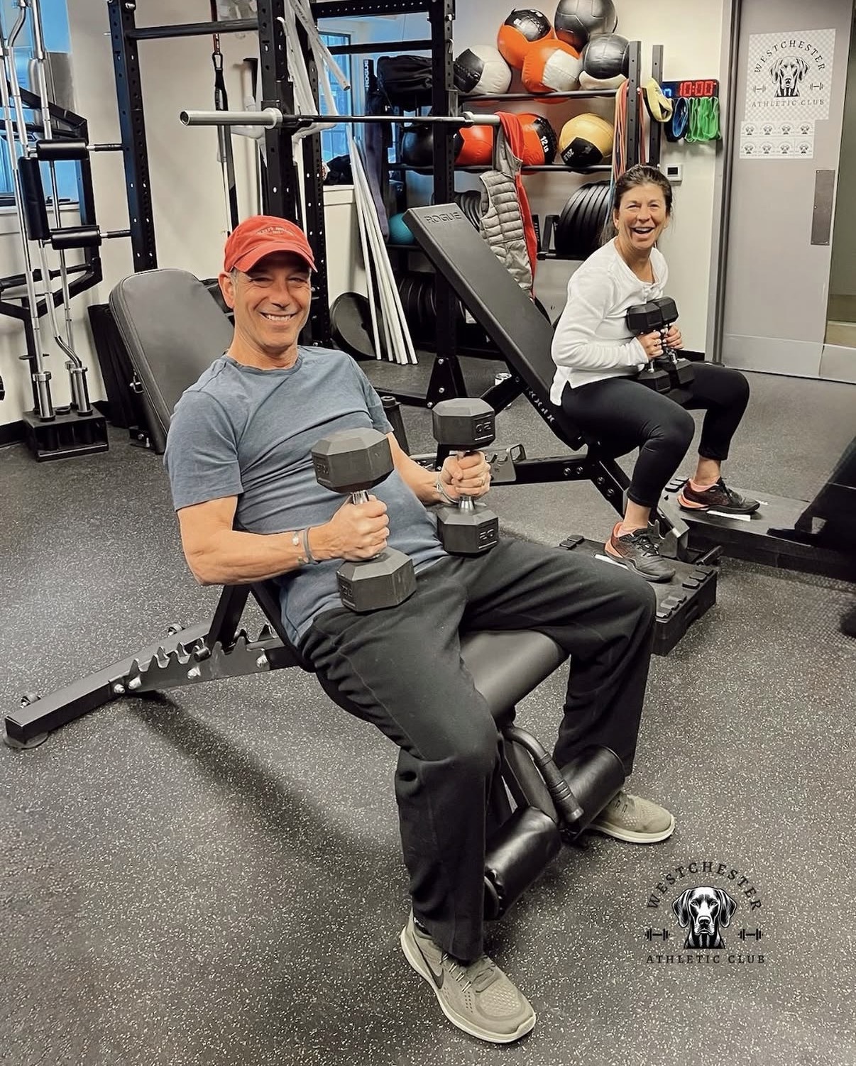 Two smiling members doing dumbbell work on adjustable benches at Westchester Athletic Club